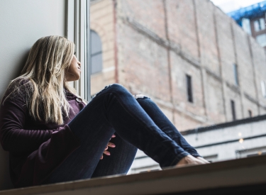A woman with long blonde hair, wearing a purple top and jeans, looks out her window at brick neighborhood buildings. Photo by Scott Warman on Unsplash