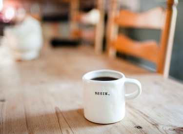 A white coffee mug that says "Begin" sits on a wooden table