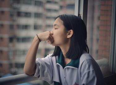 A young woman with short dark hair leans her fist on her chin and looks out a high-rise window