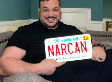 Stephen sitting on the couch and holding up a license plate that spells "NARCAN"