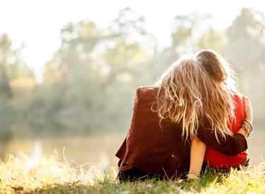 Two girls hugging while sitting in a field and facing away from the camera