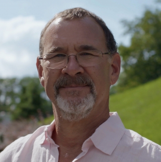 Headshot of Dr. Chudacoff: A white man with a grey beard and glasses, in a pink button down shirt, standing outside