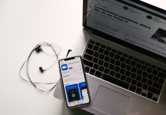 An iPhone displaying the meeting app Zoom, with headphones and a laptop. Photo by Allie Smith for Unsplash