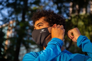 A young man with curly hair in a blue long sleeved tee puts on his face mask