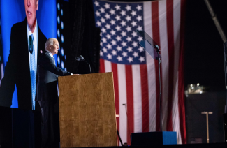 President Joe Biden giving a speech at a podium, with a large monitor and American flag in the background. Photo by Adam Schultz.